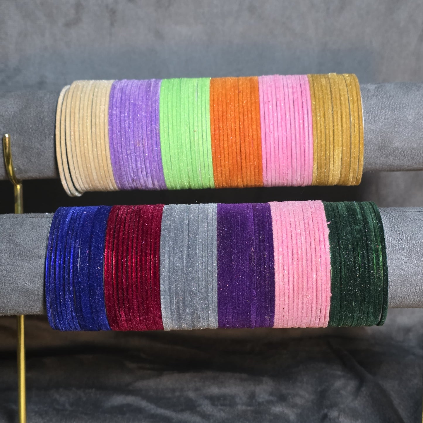"Display stand showing two rows of colorful velvet metal bangles. Top row features beige, purple, neon green, orange, pink, and mustard. Bottom row features royal blue, maroon, silver, dark purple, and bottle green bangles."