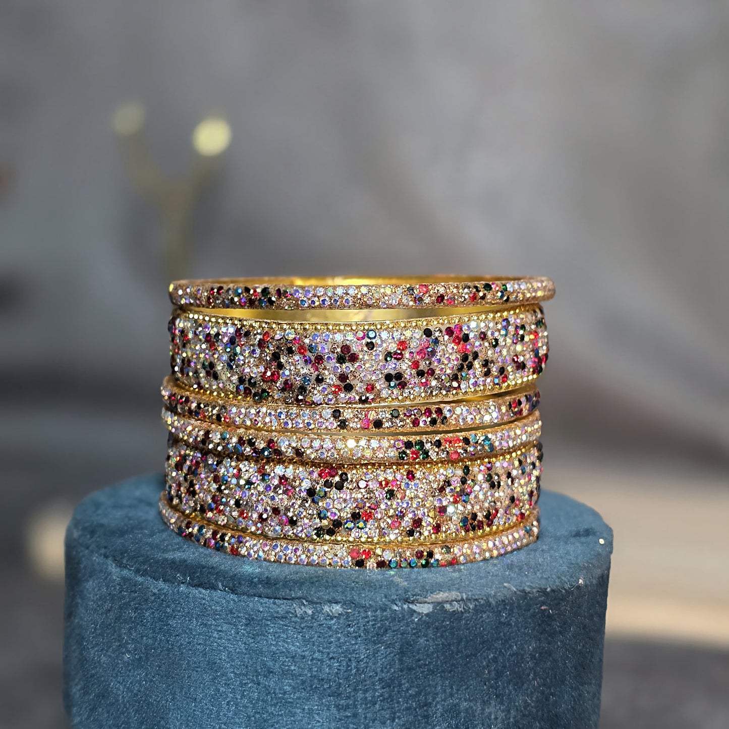 "Stack of gold-toned party wear bangles heavily encrusted with multicolored rhinestones and crystals, displayed on a blue velvet stand."