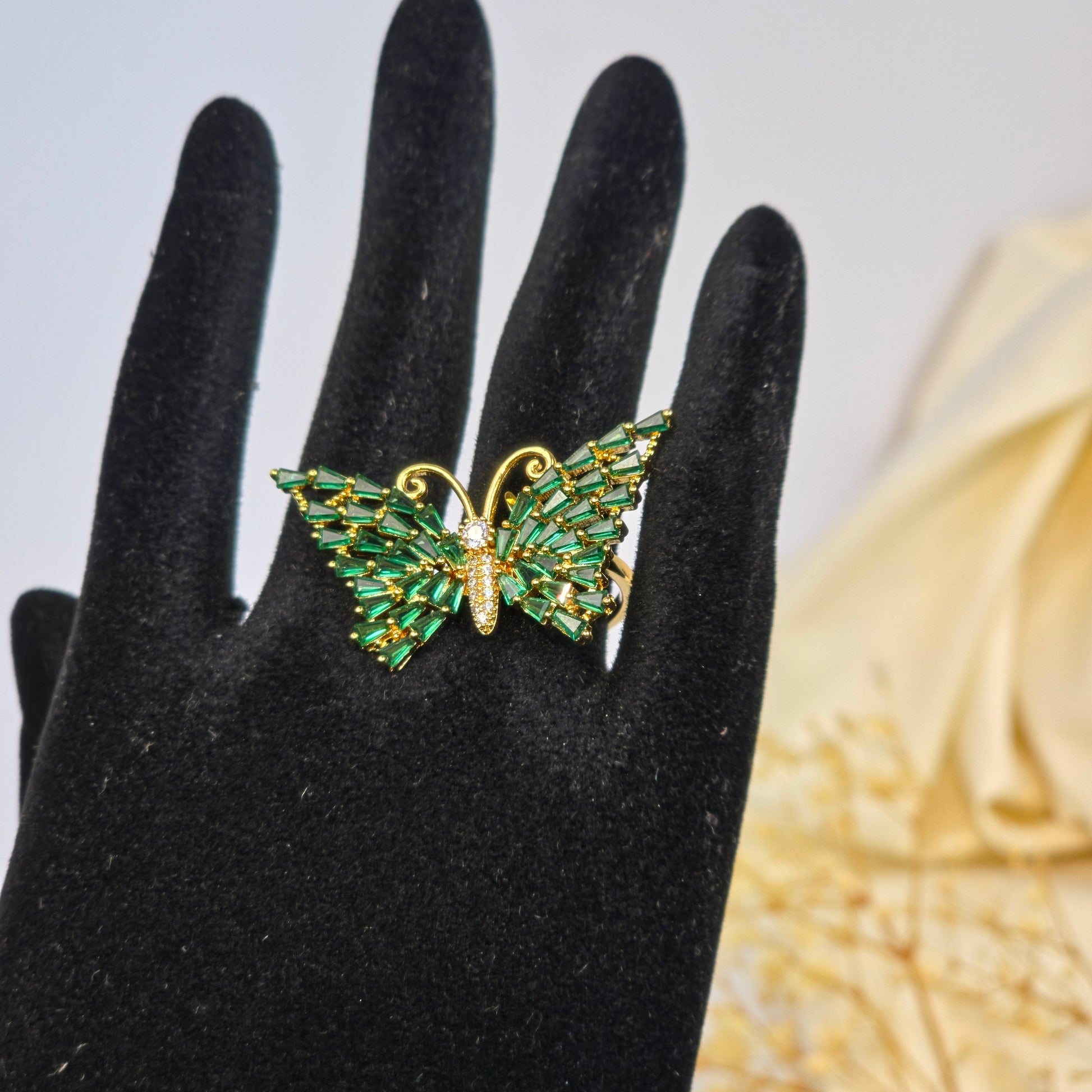 Gold and green leaf-shaped ring on a black-gloved hand with a textured gold object in the background.