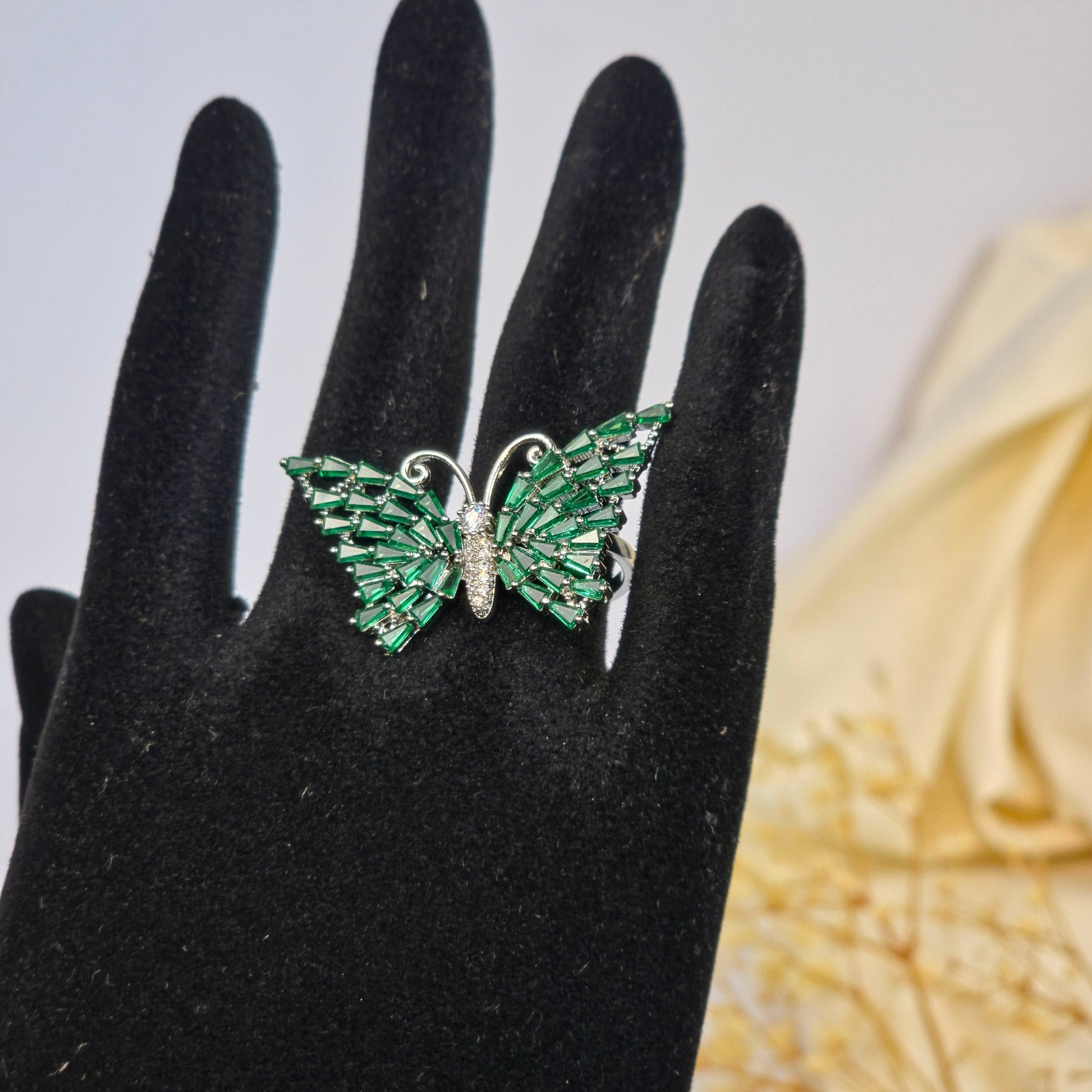 Green gemstone ring on a black-gloved hand with a blurred decorative background