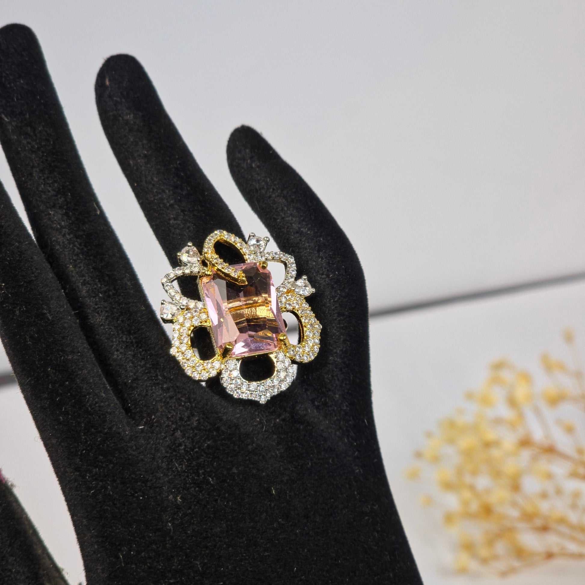 Decorative ring with pink gemstone and gold accents on a black glove against a light gray background